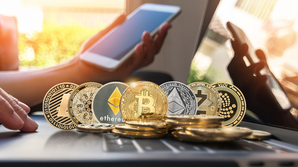 A person using a smartphone with various cryptocurrency coins like Bitcoin, Ethereum, and Cardano displayed in the foreground on a laptop, symbolizing the ease of managing digital assets on-the-go and the integration of cryptocurrency with modern mobile technology.